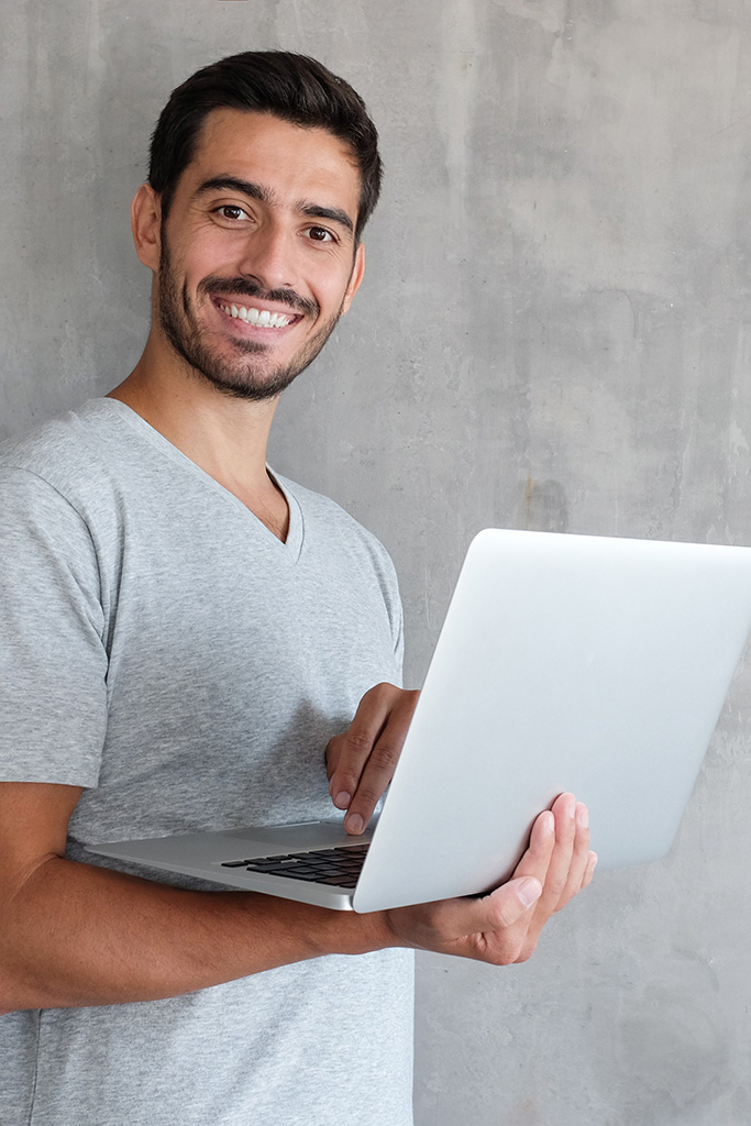 Indoor-Porträt von jungem Mann in grauem T-shirt der Laptop hält und positiv in die Kamera blickt